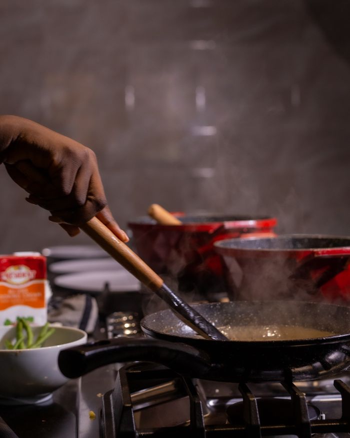 Services Chef's hand stirring a frying pan on a stove in a kitchen setting with dishes and food preparation ingredients.